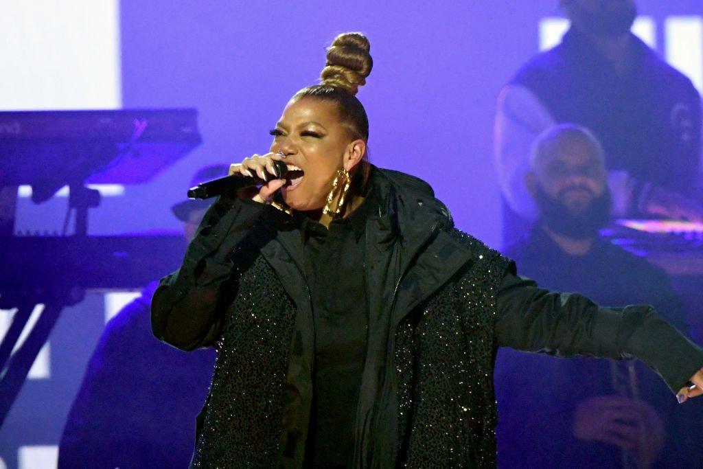 Photo of Queen Latifah performing onstage during the 65th GRAMMY Awards in 2023. She is wearing a black shirt and black jacket with gold hoop earrings and a tall bun in her hair.